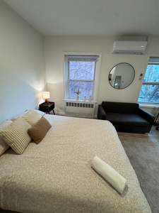 Unwind in your serene bedroom with modern comfort, featuring a cozy reading chair and peaceful tree views.