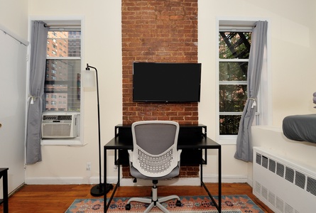Your dedicated workspace features exposed brick charm, modern desk setup, and city views through bright windows.