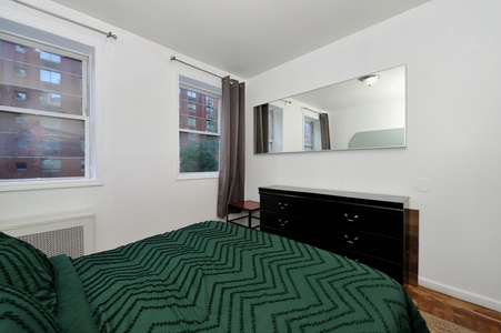 Unwind in your cozy bedroom with rich green bedding, elegant dresser, and peaceful city views through bright corner windows.