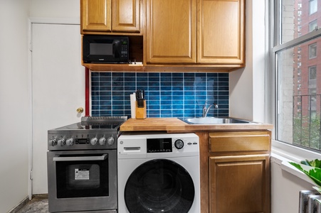 Your compact kitchen combines cooking and laundry convenience, featuring warm wood cabinets, blue tile backsplash, and city views from the window.