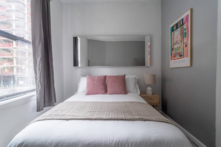 Your cozy bedroom features crisp white linens, soft pink accent pillows, and a large window offering city views to brighten your mornings.