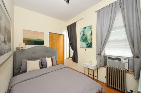 Relax in your elegant bedroom featuring a plush gray upholstered bed, original hardwood floors, and calming artwork that creates the perfect retreat.