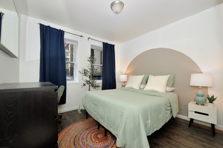 Unwind in your peaceful bedroom with soft sage linens, warm lighting, and navy curtains framing city views.