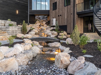 Contemporary property exterior with a dramatic multi-tiered water feature cascading through natural stone landscaping, illuminated for evening ambiance.