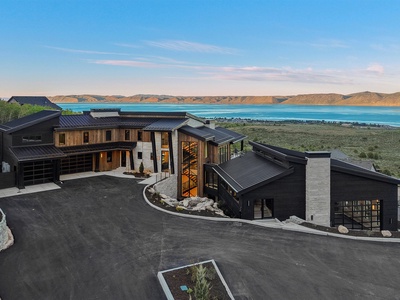 Modern architectural estate perched above turquoise lake waters, framed by rolling golden hills under a vast blue sky at day's end.