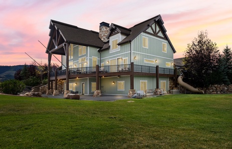 A spacious custom-built home set on manicured grounds with mountain surroundings, photographed at sunset with warm evening light illuminating the property's distinctive architecture and outdoor features.