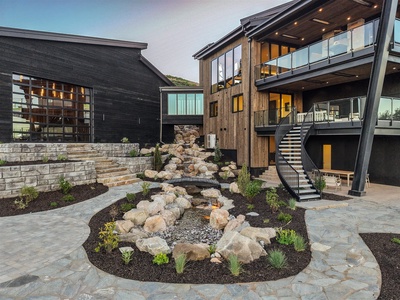 Contemporary multi-level property featuring natural wood and stone architecture with landscaped rock garden courtyard and modern outdoor lighting at dusk.