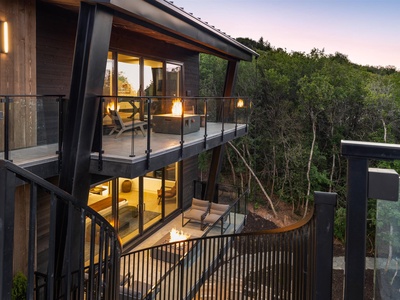 Modern multi-level architecture nestled in lush wooded hillside, featuring expansive glass walls, private balconies, and contemporary design surrounded by natural landscape.