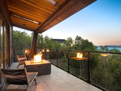 Modern covered terrace features a cozy fire table and sleek seating, set against a stunning natural backdrop where lush greenery meets water views at twilight.