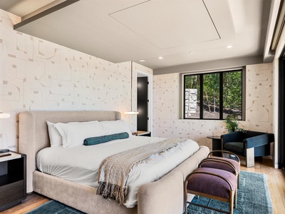 Settle into your plush bed beneath a vaulted ceiling, where soft light and artistic wallpaper create a serene retreat. Floor-to-ceiling windows frame lush forest views, inviting peaceful mornings into your private space.