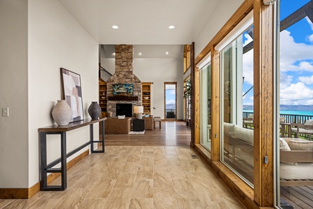 Sunlight spills through floor-to-ceiling glass doors, framing stunning blue water views that stretch to the horizon. Warm wood accents and a stone fireplace create an inviting space where you can relax and soak in the scenery.