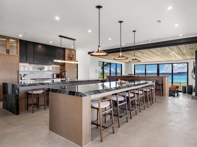 Gather around the stunning marble island while gazing at breathtaking ocean views through floor-to-ceiling windows, with elegant pendant lighting and a beautifully crafted wood ceiling creating your perfect culinary retreat.