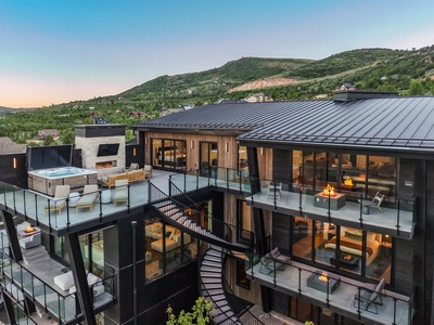 A stunning modern mountain retreat featuring sleek architecture with expansive glass walls, outdoor hot tub, and multiple balconies overlooking lush hillside scenery—the perfect setting for your alpine escape.