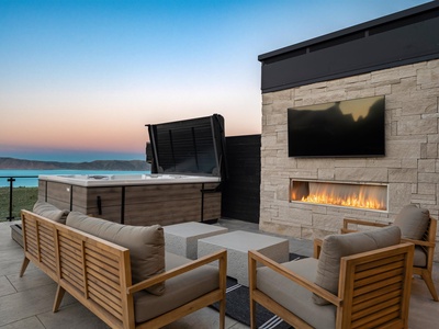 Outdoor terrace with modern stone fireplace, hot tub, and seating overlooking lake and mountain views at dusk.