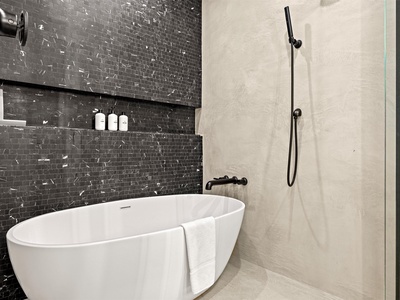 Unwind in your elegant bathroom featuring a sleek soaking tub and spacious rainfall shower. Dark textured tile contrasts beautifully with bright fixtures, creating your personal spa sanctuary.