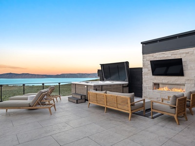 A stunning outdoor living space features an elegant stone fireplace, hot tub, and comfortable lounge seating, all set against breathtaking water and mountain views at sunset.