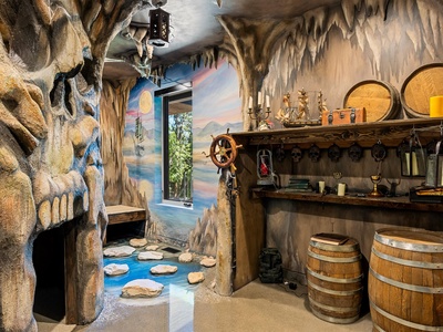 This fantastical themed interior features cave-like sculpted walls with dramatic stalactites, a painted nautical mural, ship's wheel, treasure chest, barrels, and lantern creating an immersive pirate or adventure atmosphere.