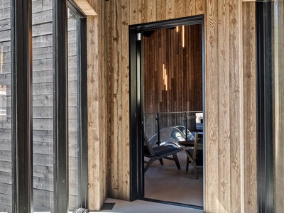 A striking modern entrance features sleek wood paneling and black-framed glass doors opening to a welcoming interior with contemporary seating and warm ambient lighting.