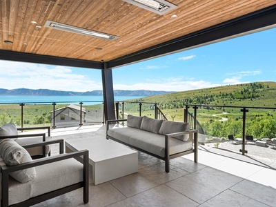 A covered outdoor terrace framed by natural wood and sleek modern lines, overlooking serene lake waters and rolling green hills stretching into the distance.