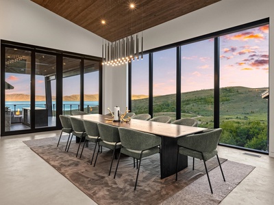 Floor-to-ceiling windows frame sweeping lake and hillside views in this modern dining space, where you'll gather around a stunning wood table beneath artistic lighting as the sky shifts to sunset hues.