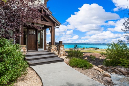 The property's entrance features stone accents and leads to stunning lakeside surroundings with expansive blue water views framed by lush landscaping.