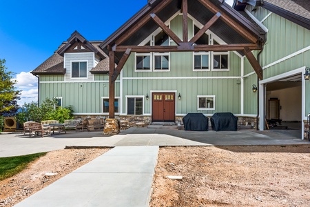 This stunning mountain lodge features distinctive timber frame architecture with sage green siding and stone accents, set against a backdrop of natural mountain terrain and expansive blue skies.