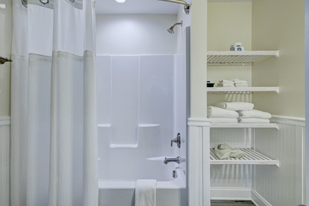 Your pristine bathroom features a spacious tub-shower combo with fresh linens organized on convenient shelving for ultimate comfort.