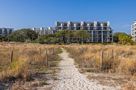 Sandy pathway winds through coastal dunes toward modern beachfront buildings, creating a scenic approach through natural landscape.