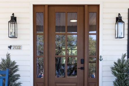A warm, welcoming entrance with a classic wood-and-glass door flanked by charming lanterns, framed by coastal greenery and nautical accents.
