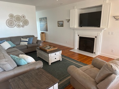 Sink into plush sectional seating by your cozy fireplace, where coastal decor and warm wood floors create the perfect retreat for relaxing evenings.