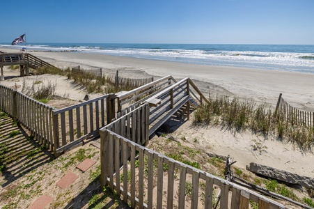 Wooden boardwalk leads through dunes to pristine beach with gentle waves and sailboat on horizon under clear blue sky.