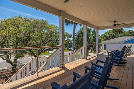 A spacious covered porch overlooking lush tropical palms and mature oaks features a row of inviting navy Adirondack chairs perfect for morning coffee or sunset relaxation.