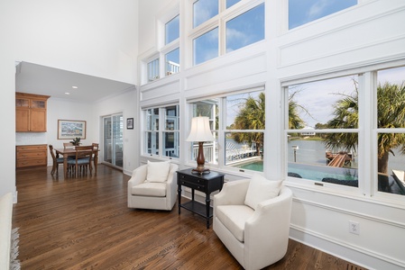 Sunlight pours through floor-to-ceiling windows, framing serene waterfront views and swaying palms. Relax in plush seating or gather at the dining table—this bright, open space invites you to unwind in coastal comfort.