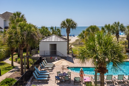Elevated view of a beachfront property featuring a swimming pool and tropical landscaping with palm trees. The main building overlooks a sandy beach and ocean beyond, with communal lounging areas.