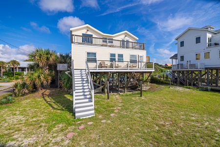 A classic coastal vacation home elevated on stilts with spacious balconies overlooking the surrounding neighborhood, set among palm trees and lush coastal vegetation under bright blue skies.