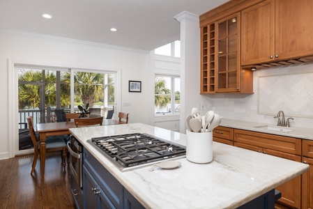 Picture yourself preparing meals in this bright, open kitchen with warm wood cabinetry, marble countertops, and a gas cooktop island. Natural light and palm tree views create your perfect coastal cooking retreat.