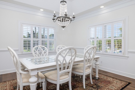 Gather around this elegant dining table, where coastal light pours through plantation shutters and a statement chandelier sets the mood for memorable meals with loved ones.
