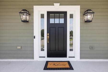 This welcoming entrance features a classic black door framed by white trim and elegant lanterns, marking the start of your coastal getaway with a warm 'Welcome' mat.