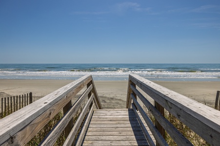 Wooden boardwalk leads to pristine sandy beach with rolling waves under clear blue skies.