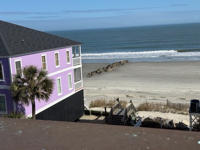 A charming purple beach house sits directly on pristine sandy shores with rolling waves and ocean views stretching to the horizon.