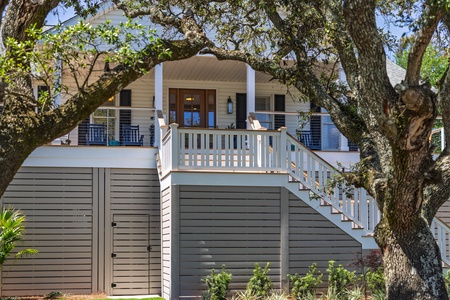 A charming coastal cottage with a covered porch and white railings, nestled among mature oak trees in a quiet neighborhood setting.