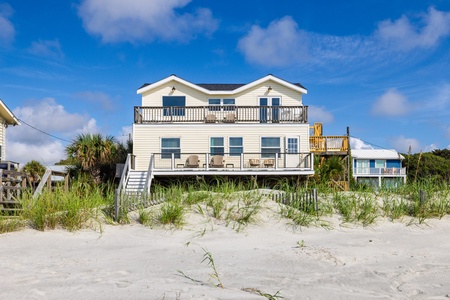 A beachfront vacation home stands directly on the sand, featuring multiple decks and immediate beach access under a bright blue sky.