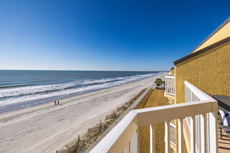 Beachfront property balcony overlooking pristine sandy shores and endless ocean views under clear blue skies.
