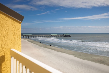 Oceanfront balcony overlooking pristine beach with historic pier extending into sparkling waters under blue skies.