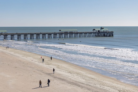 Pristine coastal area features sandy shoreline with a historic wooden fishing pier extending into rolling waves under clear skies.