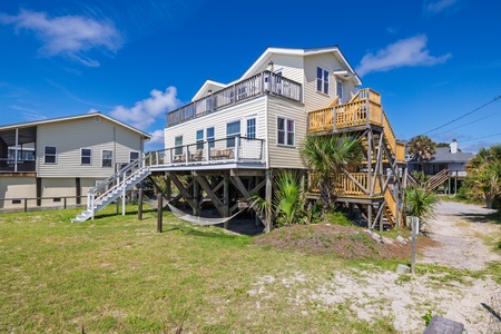 A spacious coastal home elevated on stilts, featuring multiple decks and balconies where you can enjoy the ocean breeze and surrounding views. The palm trees and sandy yard create a classic beach retreat atmosphere.