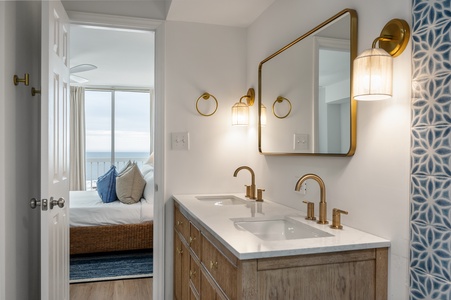 Your elegant dual-sink vanity with warm brass fixtures creates a luxurious morning routine, while ocean views beckon from your private retreat beyond.