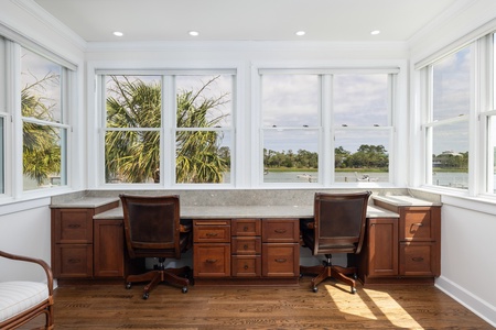 Your private workspace overlooks serene waterfront views with natural light streaming through expansive windows. The custom double-desk setup features rich wood cabinetry and comfortable seating, creating an inspiring environment for both work and relaxat