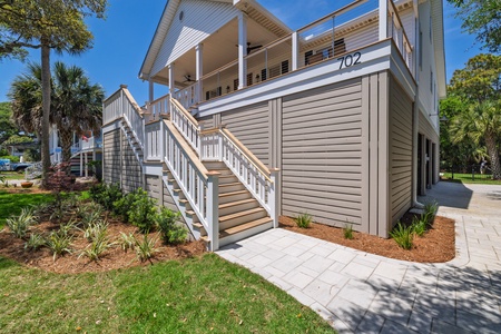 A coastal-style raised home with inviting stairs leading to a covered porch, surrounded by tropical landscaping and marked with house number 702, located in a peaceful neighborhood setting.