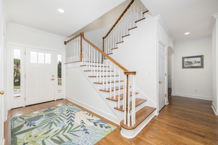 Step into your bright and welcoming entryway, where warm wood floors and a classic staircase greet you. Natural light pours through the front door's glass panels, creating an airy, inviting atmosphere for your coastal getaway.